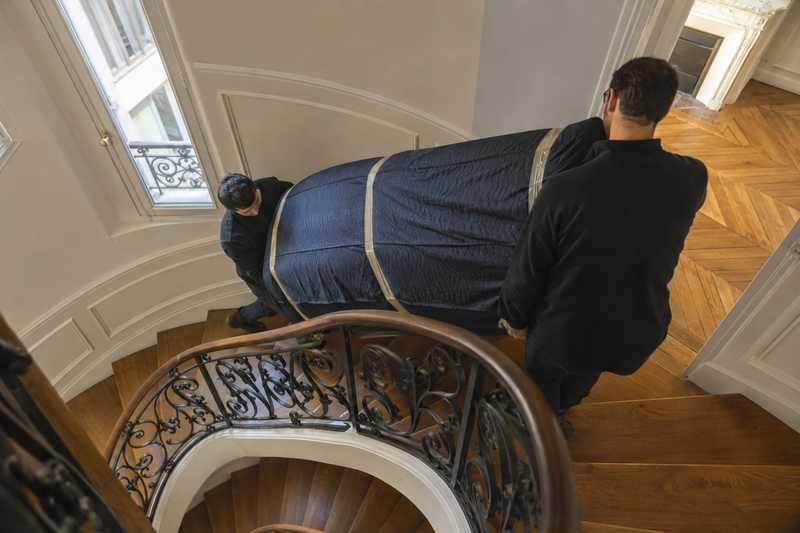 Deux demenageurs descendent un canape housse dans un escalier parisien a rampe de fer forge