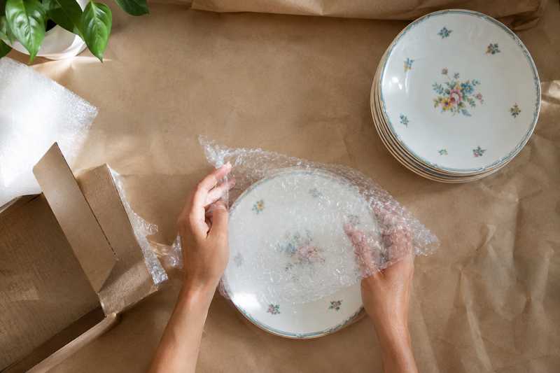 Deux mains enveloppant une assiette en porcelaine dans du papier bulle sur une table